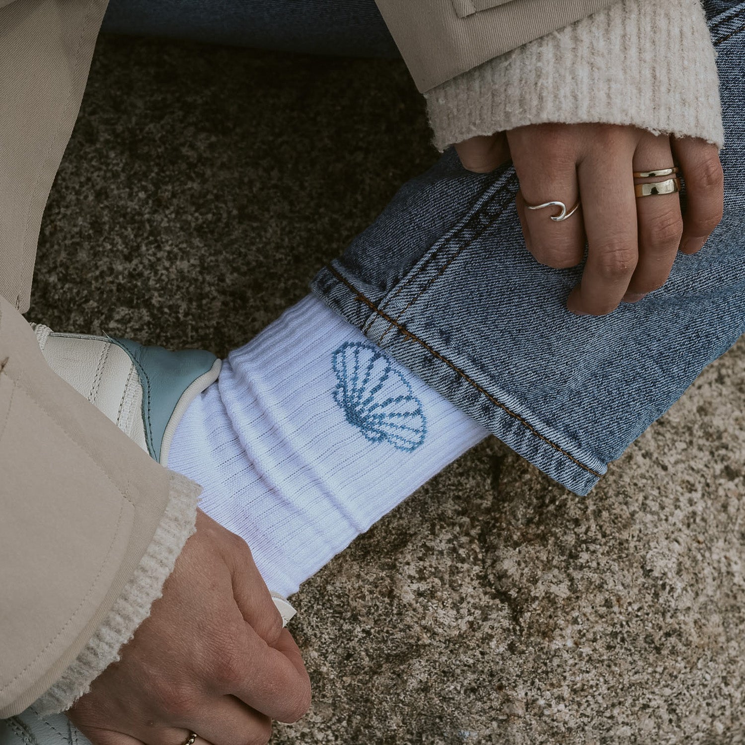 Nahaufnahme Saltwater Socken mit eingestrickter Jakobsmuschel, kombiniert mit Jeans und weißen Sneakern am Strand.