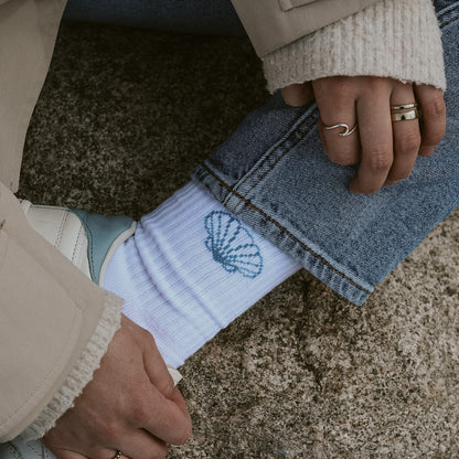 Nahaufnahme Saltwater Socken mit eingestrickter Jakobsmuschel, kombiniert mit Jeans und weißen Sneakern am Strand.