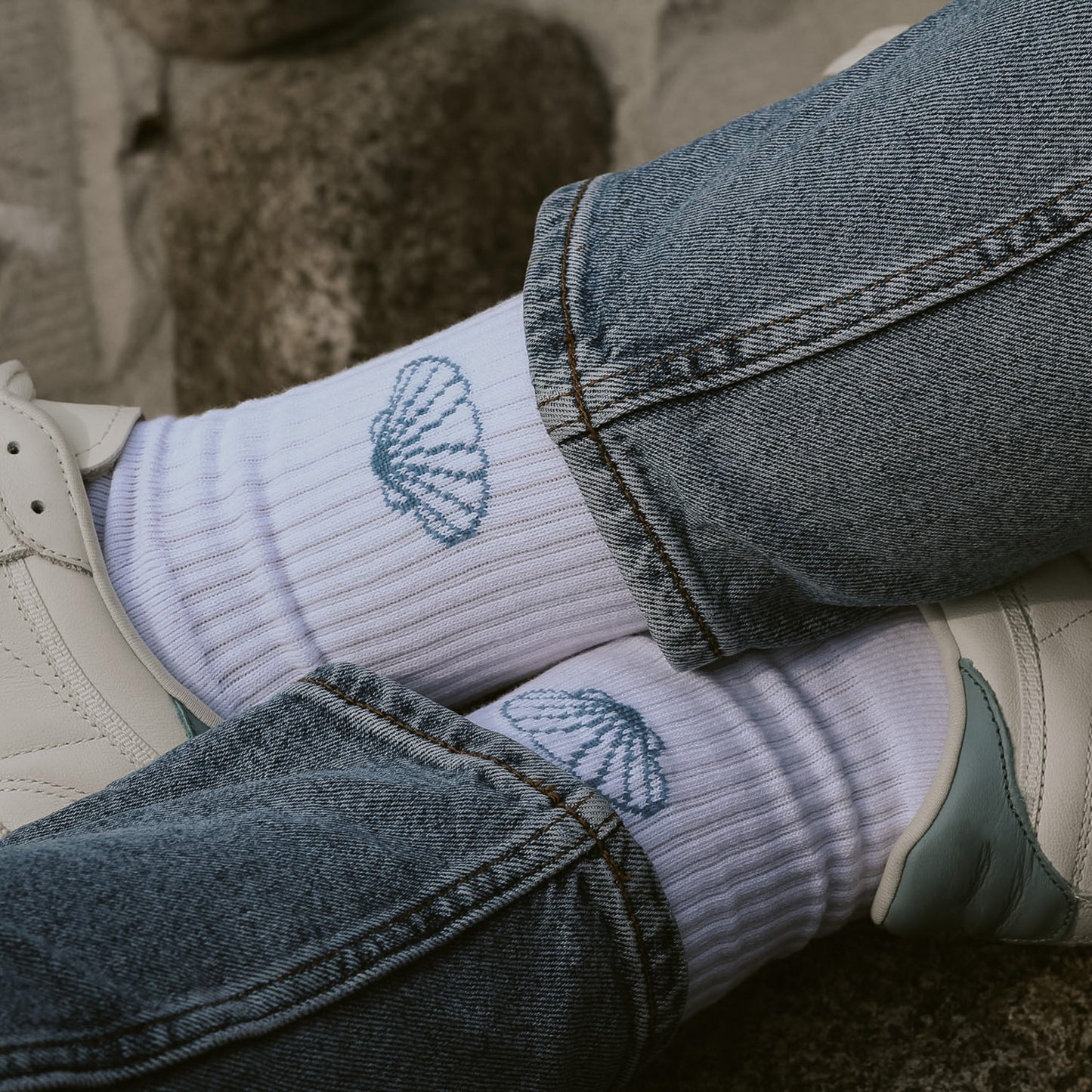 Saltwater Socken mit Muscheldesign zu Jeans und weißen Sneakern, sitzende Person auf Sandstrand. Unisex Socken in One Size Größe.