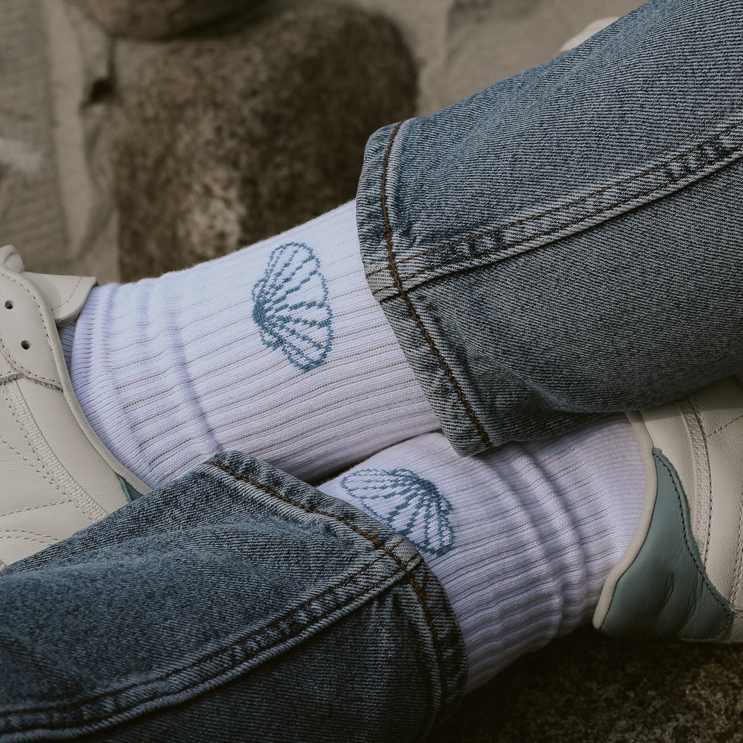 Saltwater Socken mit Muscheldesign zu Jeans und weißen Sneakern, sitzende Person auf Sandstrand. Unisex Socken in One Size Größe.