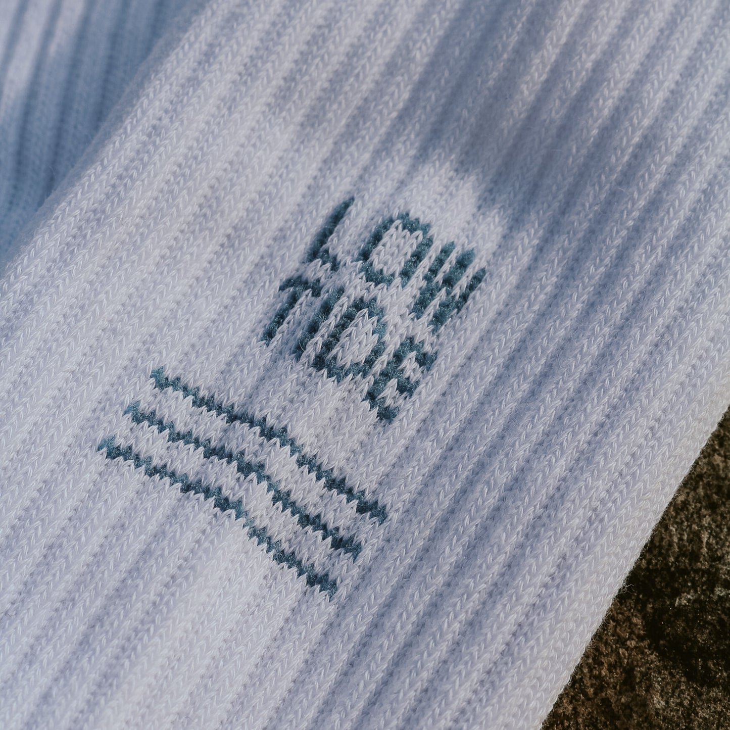 Nahaufnahme des eingestrickten Low Tide Motivs auf weißer Tennissocke – blauer Schriftzug mit flachen Wellenlinien darunter. Tennissocke von saltwaterdesigns.