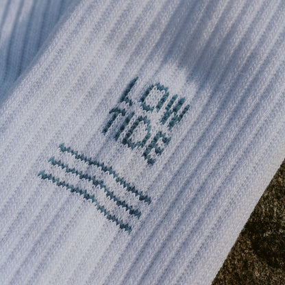 Nahaufnahme des eingestrickten Low Tide Motivs auf weißer Tennissocke – blauer Schriftzug mit flachen Wellenlinien darunter. Tennissocke von saltwaterdesigns.