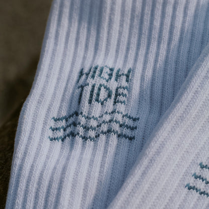Nahaufnahme des eingestrickten High Tide Motivs auf weißer Tennissocke – blauer Schriftzug mit hohen Wellenlinien darunter. Onesize Größe.