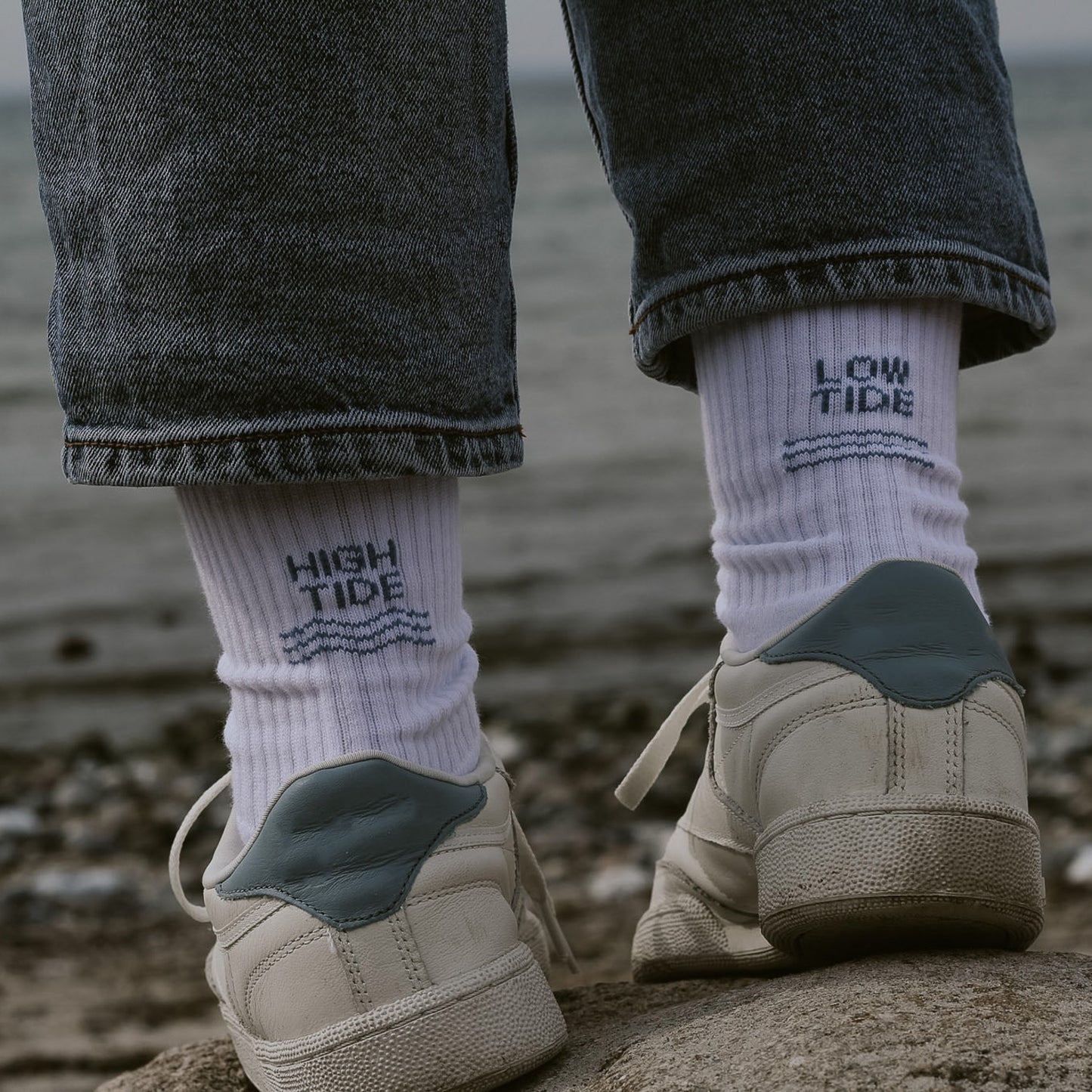 Person von hinten mit weißen Saltwaterdesigns Tennissocken aus Bio-Baumwolle, High Tide links, Low Tide rechts – getragen mit weißen Sneakern an einem Kieselstrand.