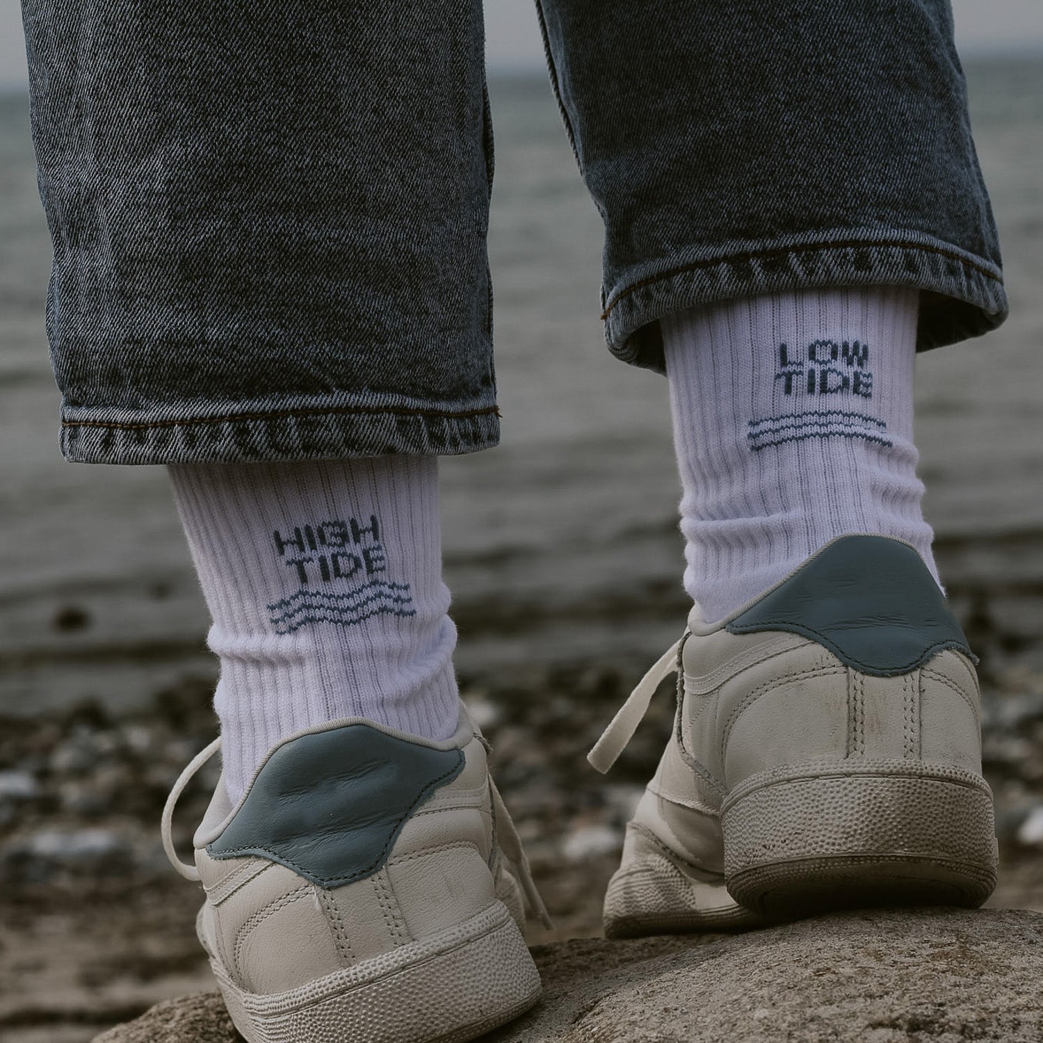 Person von hinten mit weißen Saltwaterdesigns Tennissocken aus Bio-Baumwolle, High Tide links, Low Tide rechts – getragen mit weißen Sneakern an einem Kieselstrand.