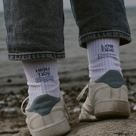 Person von hinten mit weißen Saltwaterdesigns Tennissocken aus Bio-Baumwolle, High Tide links, Low Tide rechts – getragen mit weißen Sneakern an einem Kieselstrand.