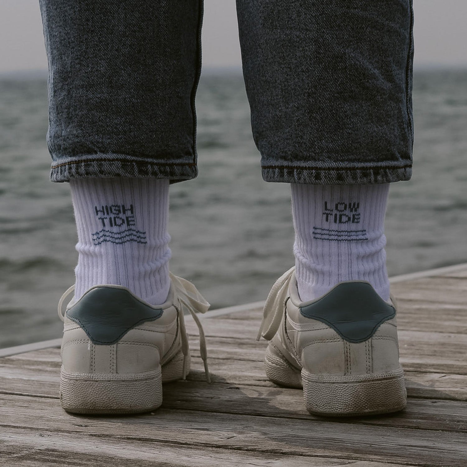 Person von hinten mit weißen Saltwaterdesigns Tennissocken, High Tide links, Low Tide rechts – getragen mit weißen Sneakern auf einem Holzsteg mit Meer im Hintergrund.