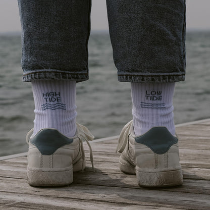 Person von hinten mit weißen Saltwaterdesigns Tennissocken, High Tide links, Low Tide rechts – getragen mit weißen Sneakern auf einem Holzsteg mit Meer im Hintergrund.