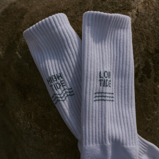 Weiße Tennissocken aus Bio-Baumwolle mit eingestricktem High Tide und Low Tide Motiv, auf einem Stein liegend – Detailansicht beider Socken von oben.