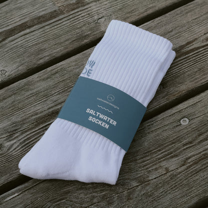 Weiße Saltwaterdesigns Tennissocke High Tide Low Tide, fertig verpackt mit der petrolblauen saltwaterdesigns Banderole, auf einem Holzuntergrund liegend.