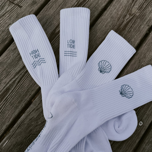 Vier weiße Saltwater Designs Tennissocken mit High Tide, Low Tide und Muschel-Motiv, locker übereinander auf einem Holzuntergrund liegend – Detailansicht aller eingestrickten Designs.
Von saltwaterdesigns.