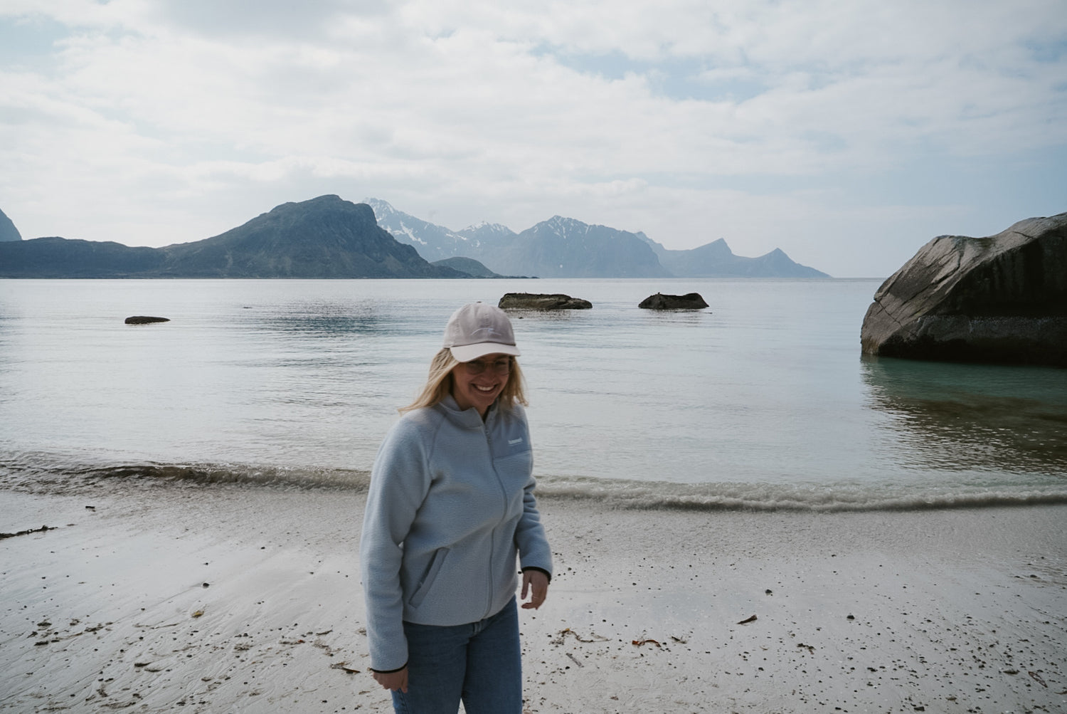 Frau mit hellblauer Jacke und Cap am Strand in Norwegen, im Hintergrund Meer und schneebedeckte Berge